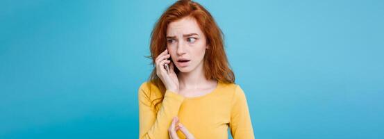 Lifestyle concept - Portrait of ginger red hair girl with shocking and stressful expression while talking with friend by mobile phone. Isolated on Blue Pastel Background. Copy space photo
