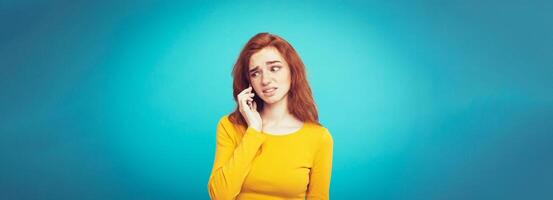 Lifestyle concept - Portrait of ginger red hair girl with shocking and stressful expression while talking with friend by mobile phone. Isolated on Blue Pastel Background. Copy space photo