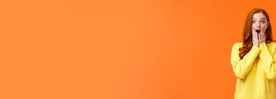 Impressed and speechless redhead woman found out awesome news, gasping, drop jaw astonished, touch cheeks and stare startled camera, christmas shopping season started, orange background photo