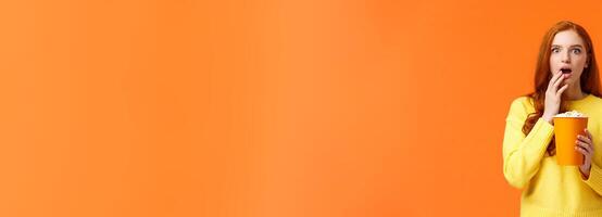 Shocked and interested, entertained redhead girl watching awesome new movie on big screen, eating popcorn, gasping open mouth astounded, startled of cool film plot, orange background photo