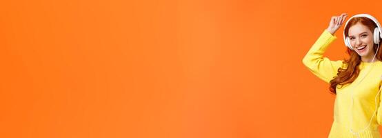 Lets dance. Cheerful carefree modern hipster girl with red curly hair and freckles, dancing lift hand up and holding smartphone, listen music with new ordered online headphones, orange background photo