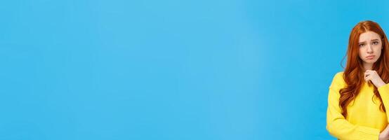Upset redhead girl showing empathy, pity for friend hearing sad news, frowning and sulking feeling uneasy for person got in trouble, standing over blue background distressed or depressed photo