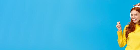 Cheeky and joyful, happy smiling redhead ecstatic woman having fun, listen music and lifting hands up as dancing, enjoying awesome sound quality, standing blue background delighted photo