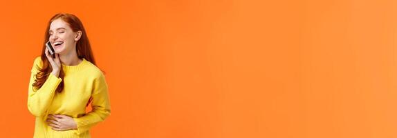 Communication, technology and people concept. Cheerful pretty redhead hipster girl talking on phone and laughing carefree, touching belly close eyes, smartphone near ear, orange background photo