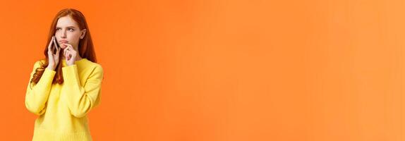 Indecisive and unsure, skeptical serious-looking redhead woman having tough decision make during conversation on phone, frowning touch lip pensive, order food delivery, orange background photo