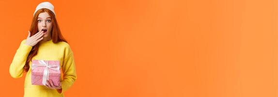 Girl surprised didnt expact gift from you. Amused and startled cute redhead girlfriend open mouth, cover it with hand as receive cute pink box of present, look camera excited, orange background photo