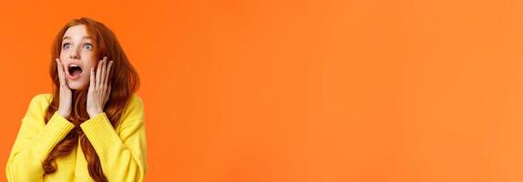 Oh no something falling from sky. Shocked and speechless, startled young concerned redhead woman drop jaw, gasping and staring upper left corner concerned, touching face worried, orange background photo