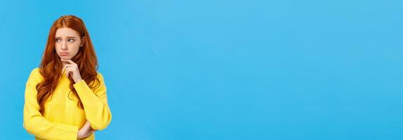 Sad timid and lonely redhead woman too feeling let down and depressed, sulking looking upset and envy left, frowning touching lip pensive, having bad feeling, standing anxious over blue background photo