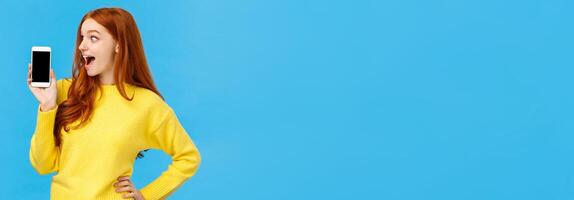 Wow look at this app. Amused and wondered, excited redhead girl looking at smartphone display, showing mobile screen, shopping site or application, standing blue background photo
