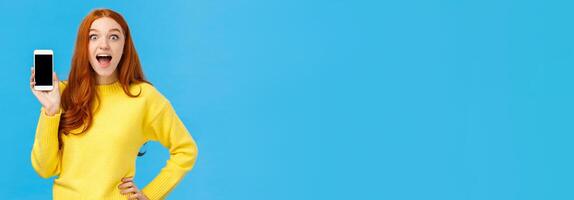 Astonished and impressed, excited redhead female in yellow sweater introduce new app, showing smartphone display, smiling fascinated open mouth amused, look camera, blue background photo