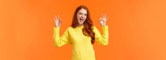Cheerful and positive, enthusiastic redhead female in yellow sweater winking in approval, smiling joyful, give permission, positive reply, rate good movie, nod agreement, recommend, orange background photo