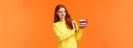 No thanks, dont want. Cute redhead girl trying resist temptation take bite delicious sweets, holding tasty cake and showing stop, refusal or rejection with grimacing face, express aversion, dislike photo