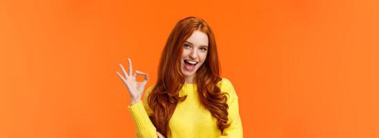 Sassy and daring cool redhead girl with curls, showing okay excellent gesture and smiling excited, assure party be awesome, give permission, say yes, confirm or recommend something, orange background photo