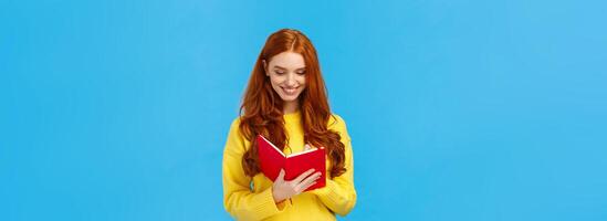 Waist-up portrait redhead female student write down thoughts in red lovely notebook, prepare grocery list, make schedule for college lectures, smiling as sharing thoughts in diary, blue background photo