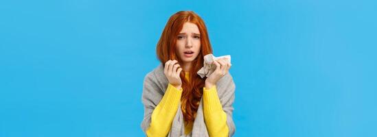 Dizzy, uneasy sad redhead female wearing scarf to get warm, catching cold, sneezing and having runny nose, using napkin, looking camera with tired exhausted expression, feel fatigue, blue background photo