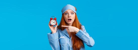 Shocked and concerned, worried cute redhead girl pointing at alarm clock with frustrated, nervous expression, being late, dont know how set up right time, standing blue background in nightwear photo