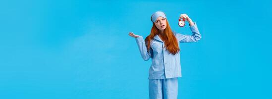 Why didnt you set alarm. Confused and unsure upset attractive redhead woman in pyjama and sleep mask, shrugging perplexed staring questioned camera, holding clokc, blue background photo