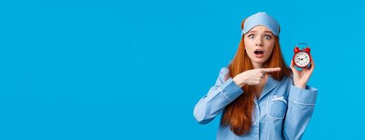 Shocked and concerned, worried cute redhead girl pointing at alarm clock with frustrated, nervous expression, being late, dont know how set up right time, standing blue background in nightwear photo