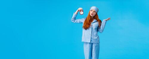 Why didnt you set alarm. Confused and unsure upset attractive redhead woman in pyjama and sleep mask, shrugging perplexed staring questioned camera, holding clokc, blue background photo