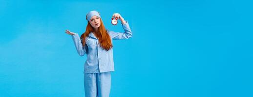 Why didnt you set alarm. Confused and unsure upset attractive redhead woman in pyjama and sleep mask, shrugging perplexed staring questioned camera, holding clokc, blue background photo