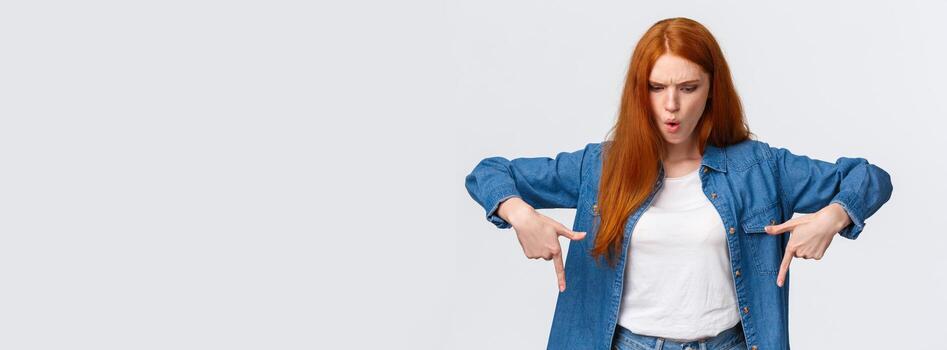 Wait a minute what is that. Frustrated and angry tensed redhead girlfriend asking wtf going on, dont understand, looking perplexed and mad down, pointing bottom copy space, stand white background photo