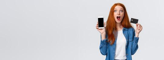 Waist-up portrait excited, astonished and thrilled redhead female student holding credit card, showing smartphone display, application, drop jaw impressed, looking camera astounded, white background photo