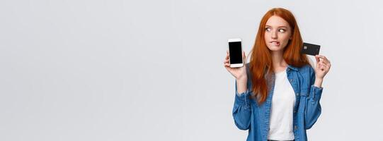 Girl consider buy thing with super good discount. Temped and wishful excited redhead female shopaholic looking at smartphone, shopping mobile app, holding credit card, bite lip eager purchase photo