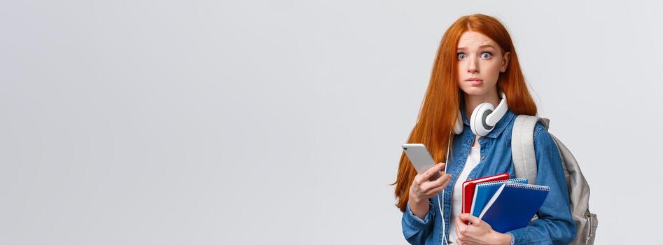 indeciso, nervioso linda pelirrojo estudiante de primer año chica, comienzo nuevo año colega, mirando preocupado cámara, mordiendo labio nerviosamente, participación mochila, cuadernos y teléfono inteligente, leyendo mensaje foto