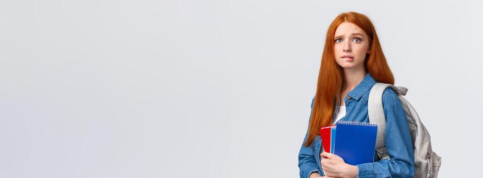 preocupado linda y nervioso pelirrojo Adolescente niña asustado padres regaño su para salto a la comba clase, mordiendo labio ansiosamente mirando cámara, participación cuadernos y mochila terminado blanco antecedentes foto