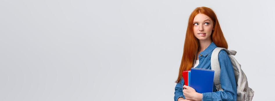 creativo y pensativo linda atrevido astuto niña con rojo cabello, sostener mochila y cuadernos, mordiendo labio mirando arriba reflexionando, querer omitir clase y tener divertido amigos, pensando terminado blanco antecedentes foto
