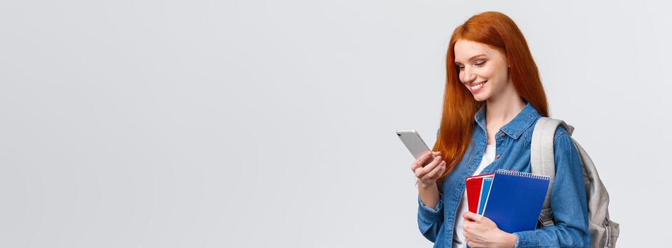 moderno estilo de vida, tecnología y educación concepto. lindo, encantador pelirrojo hembra estudiante Bóveda a instalaciones después clases en colega, participación mochila y cuadernos, chateando en teléfono inteligente foto