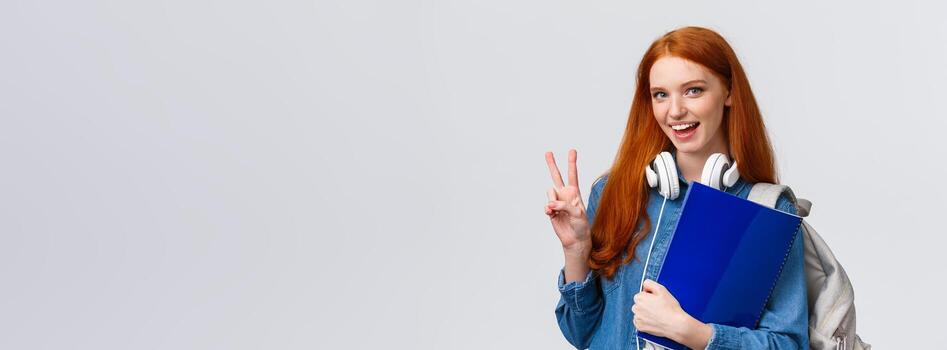 encantador simpático Universidad alumno, pelirrojo niña con documentos, documentos para escuela, participación mochila y vestir auriculares terminado cuello, hacer paz gesto informal saludo compañera de clases, blanco antecedentes foto