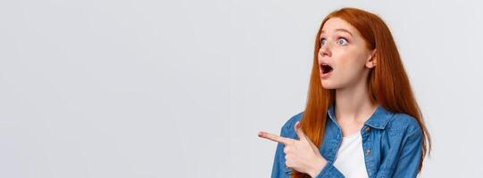 Have you seen it. Close-up astonished and impressed shook redhead girl saw something impressive, open mouth excited, pointing finger left, watching awesome performance, white background photo