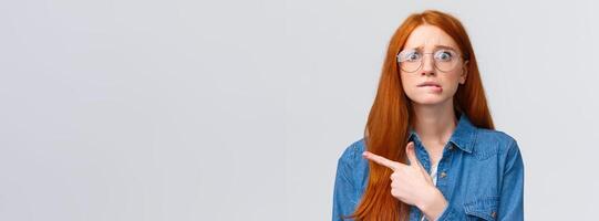 Nervousness, people and education concept. Anxious timid and insecure cute redhead girl in glasses, student worrying biting lip, hesitating telling something, pointing finger left, white background photo