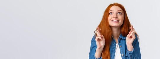 Excited and hopeful, optimistic cute redhead girl want dream come true, desire something, aspire achieve success good results, smiling looking up sky and making wish with fingers crossed good luck photo