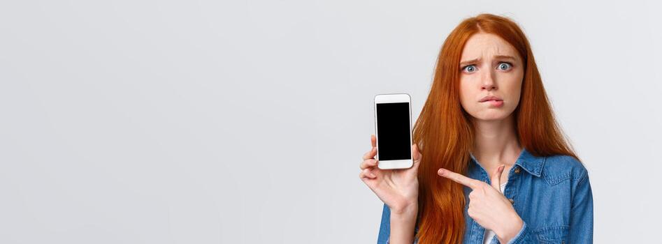 Worried, insecure and alarmed cute redhead girl asking for advice having problems messaging with stranger on dating app, dont know what answer standing nervous pointing finger at smartphone display photo