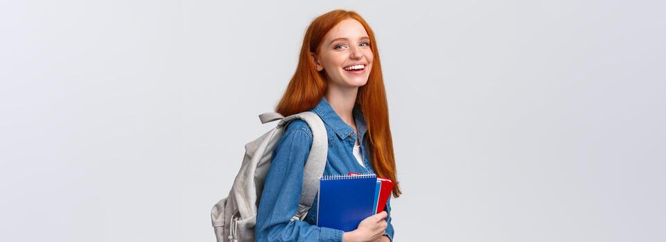 estudiantes, educación y estudiando concepto. encantador carismático pelirrojo astuto niña con mochila, cuadernos y estudiar libros, en pie blanco fondo, sonriente alegre mirando cámara foto
