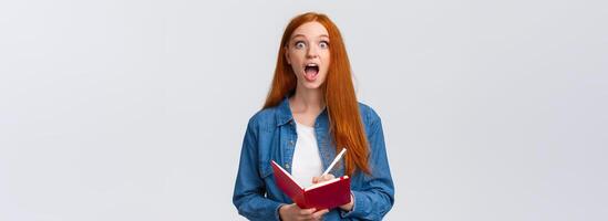 Amused and excited, astonished redhead girl fascinated with amazing lecture giving speech, writing down useful notes, holding notebook and staring thrilled camera, white background photo