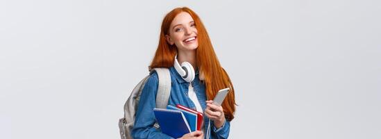 Cheerful, friendly and charismatic redhead woman laughing joyfully, texting friends, chatting with smartphone, enjoying college lifestyle, heading campus with backpack and notebooks photo