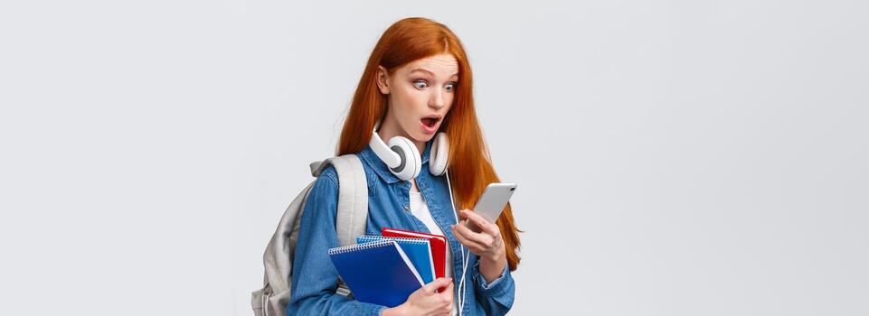 conmocionado y asombrado, jadeando Sin palabras pelirrojo hembra estudiante con mochila y cuadernos, curioso a teléfono inteligente pantalla tembló abierto boca y popular ojos móvil monitor foto