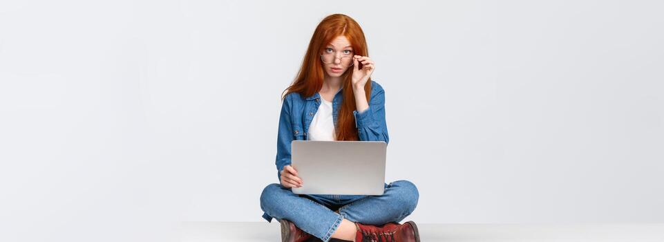 Excuse me what, I am busy. Questioned redhead female student interrupted during preparing project, searching material for class in internet, look curious camera, sitting on floor with laptop photo