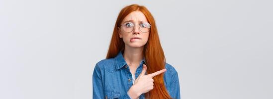 Nervousness, people and education concept. Anxious timid and insecure cute redhead girl in glasses, student worrying biting lip, hesitating telling something, pointing finger left, white background photo
