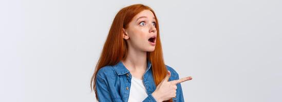 Have you seen it. Close-up astonished and impressed shook redhead girl saw something impressive, open mouth excited, pointing finger left, watching awesome performance, white background photo