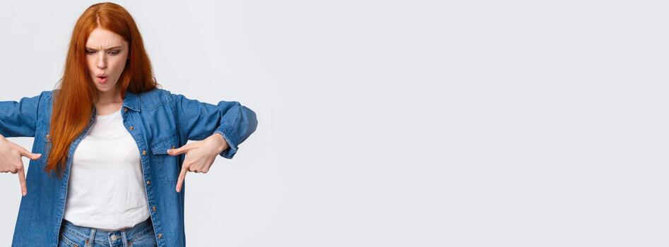 Wait a minute what is that. Frustrated and angry tensed redhead girlfriend asking wtf going on, dont understand, looking perplexed and mad down, pointing bottom copy space, stand white background photo