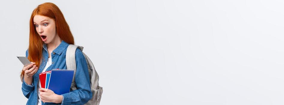 increíble No clases hoy. asombrado y emocionado pelirrojo Adolescente niña jadeando como leyendo frio mensaje en teléfono inteligente, participación mochila cuadernos y móvil teléfono, blanco antecedentes foto