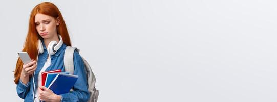 Silly sad and gloomy pouting girl with red foxy hair, frowning sighing and looking with regret smartphone screen, reading troublesome message, feeling bad, stand with backpack and notebooks photo