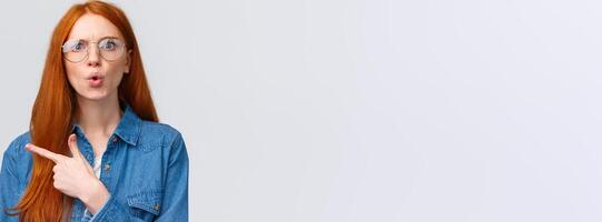Confused and shook questioned redhead woman asking question and stare shocked and displeased, pointing finger left at something bothering and strange, standing complaining over white background photo