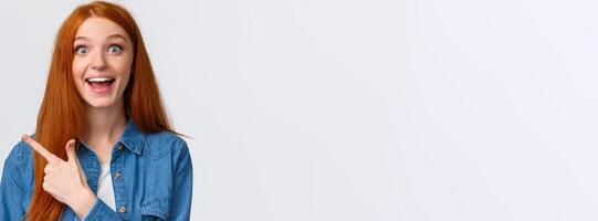 Close-up portrait excited happy ginger girl, foxy hair and blue eyes, smiling upbeat with amazement and joy, pointing finger left, attend amusing cool party, telling about it, white background photo