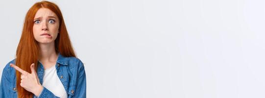 Anxious and worried concerned redhead caucasian girl having big problems, nervously biting lip and frowning scared, pointing afraid left, standing white background waiting be scolded photo