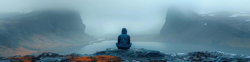 A person is seated on a rock, gazing at a lake amid mountain scenery photo
